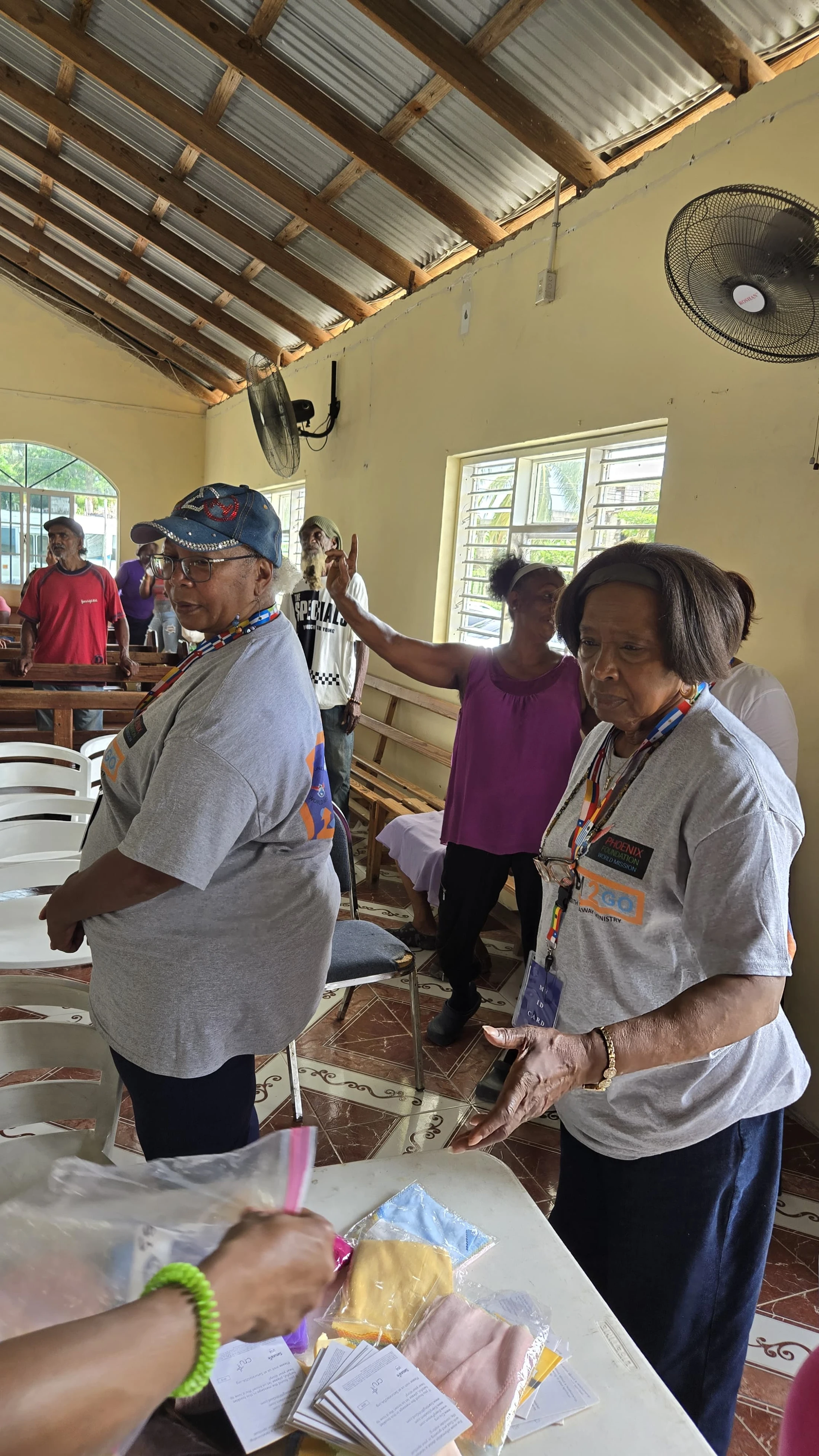 A group of people are gathered inside a simple room with a corrugated metal roof. Two older women in the foreground wear matching gray volunteer t-shirts.