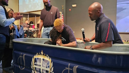 A woman with her hands clasped in prayer is being baptized in a blue portable baptistry as a pastor speaks into a microphone and others watch.