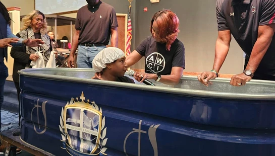 A person wearing a white cap sits in a blue baptismal font while a man and a woman perform the religious ceremony, as others watch.