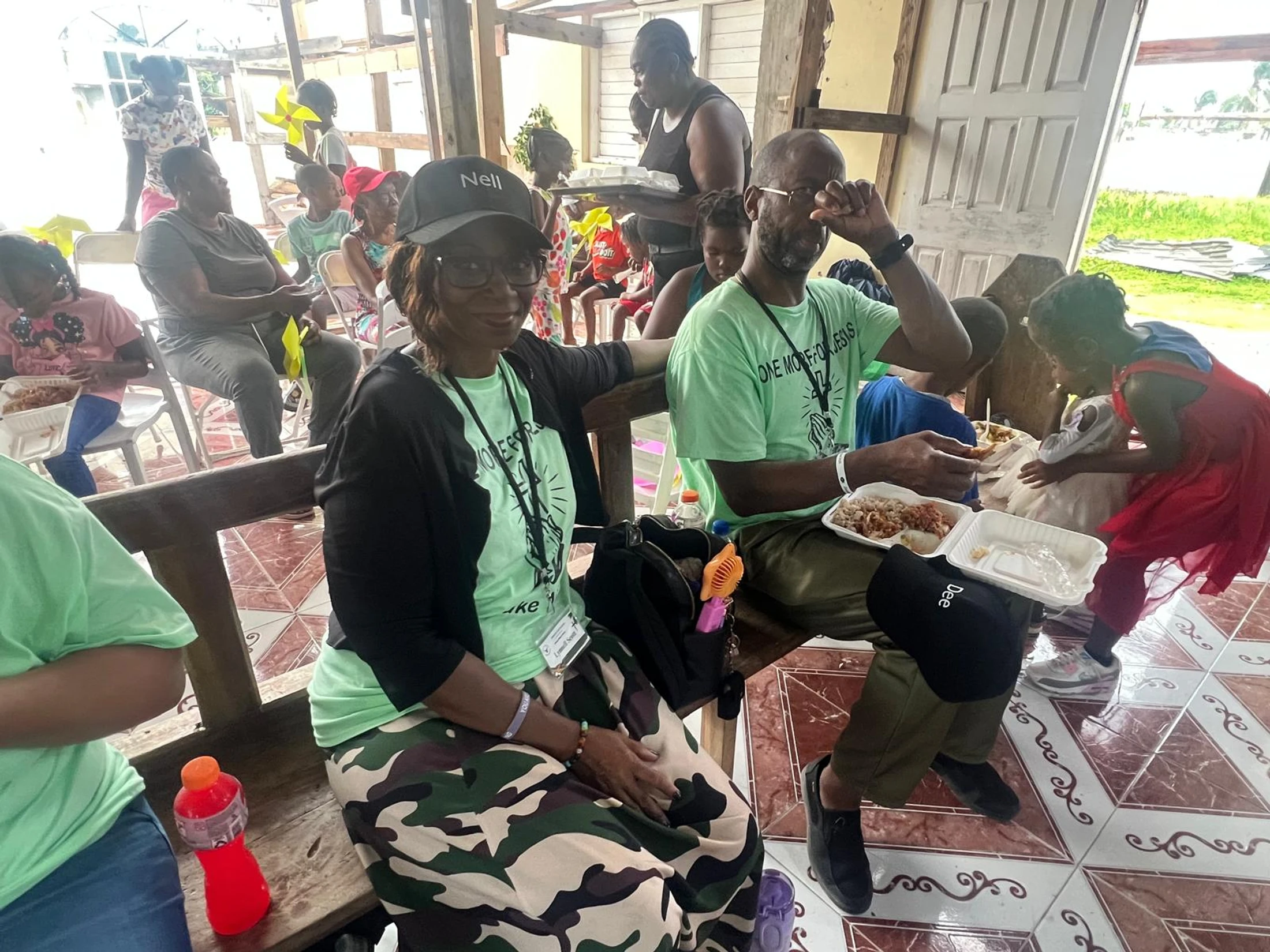 A group of adults and children gather on benches and chairs to eat a meal together. A woman in a cap and a man in a green shirt sit in the foreground.