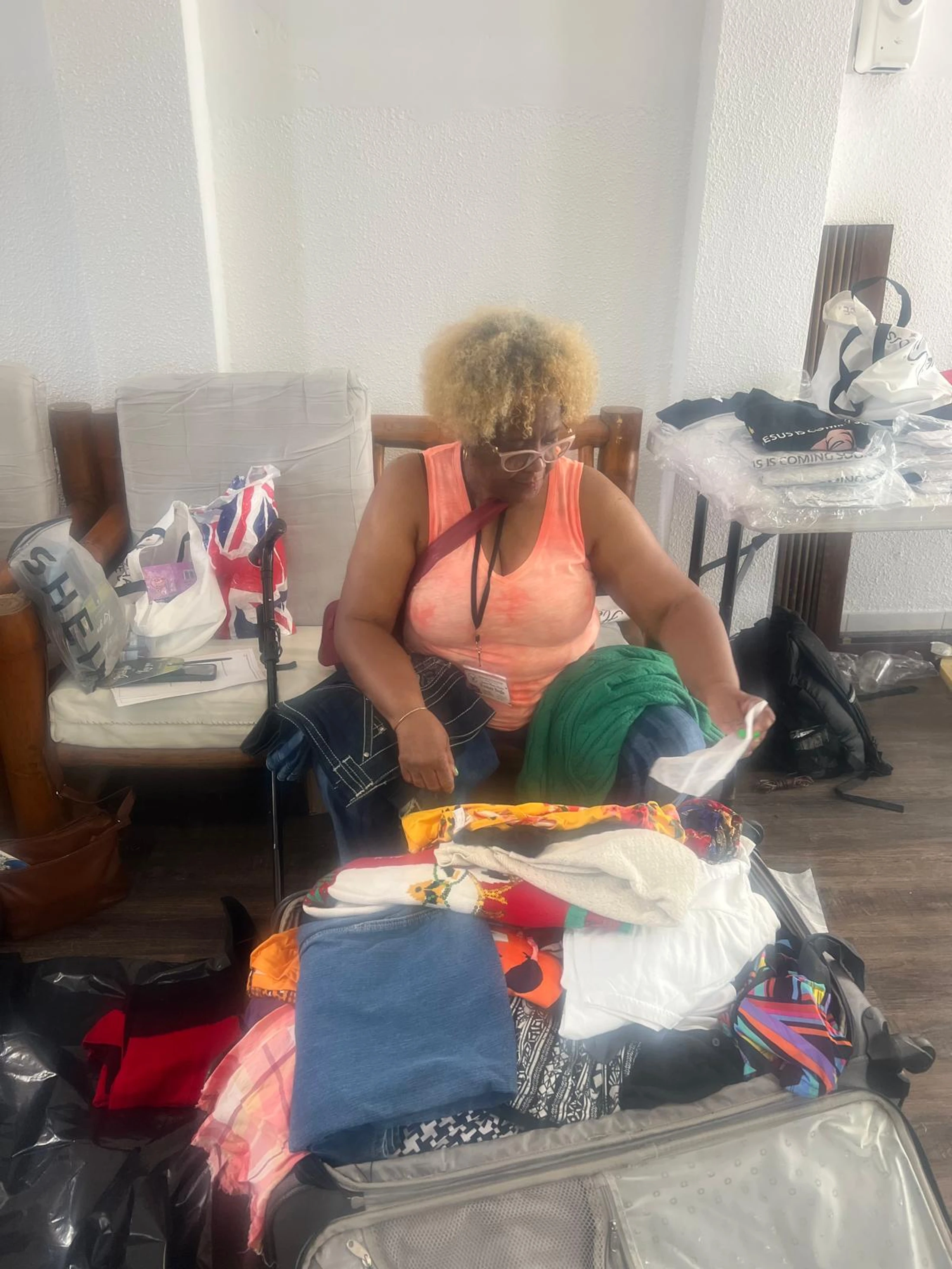 A woman with blonde hair sits in a chair and packs an overflowing suitcase on the floor with many colorful clothes and jeans.