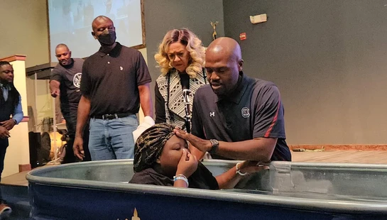 A pastor prepares to baptize a woman by full immersion in a portable baptismal font on a stage, with other church members watching the ceremony.