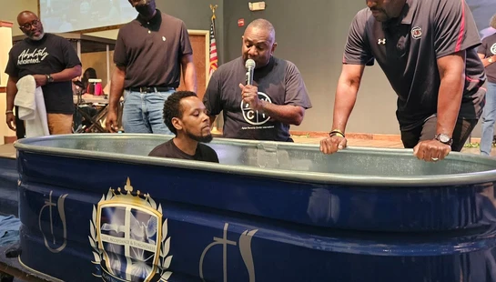 A man sits in a blue portable baptismal font on a stage, while a pastor speaks into a microphone and two other men stand by to assist in the ceremony.