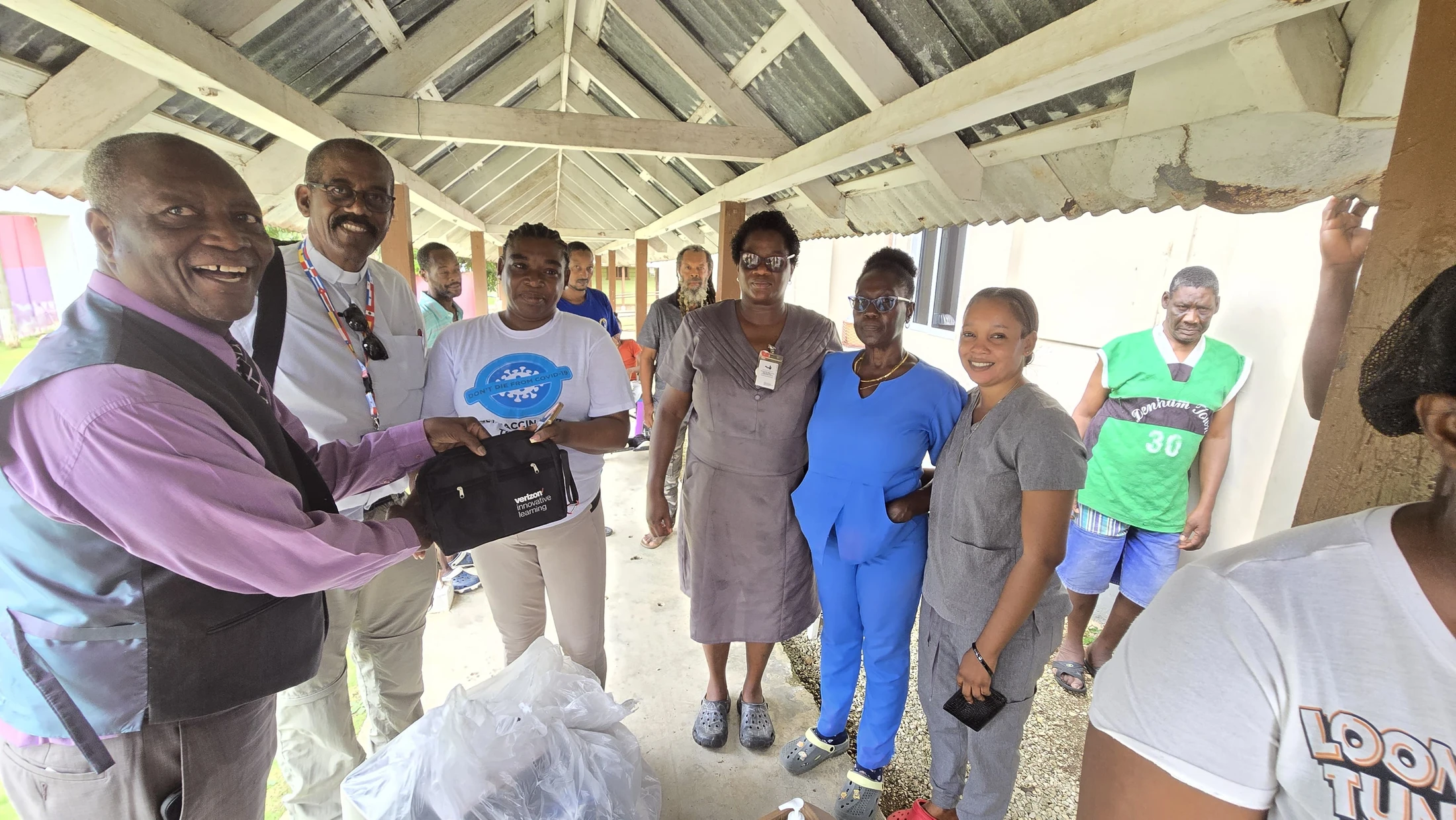 A smiling man in a vest hands a black Verizon bag to a woman as a group of people, including healthcare workers, watch under a covered walkway.
