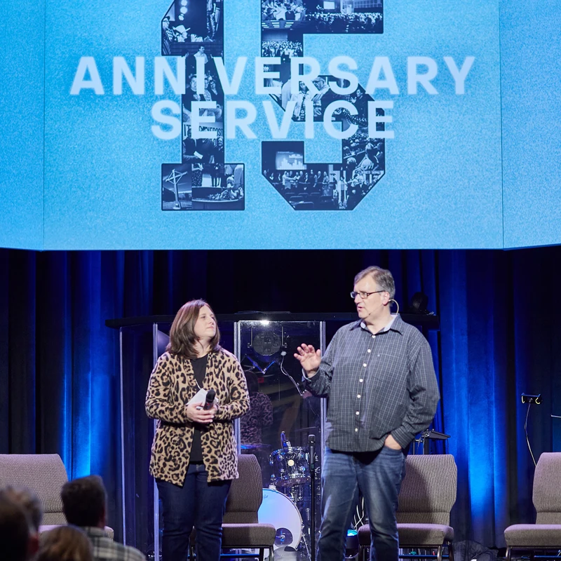 A man and a woman stand on a stage speaking to an audience, in front of a large blue screen that reads '15 Anniversary Service'.