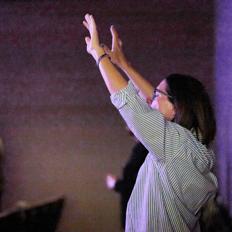 A woman with dark hair and glasses, seen in profile, raising her hands in worship. She wears a striped shirt in a room with dim, purple lighting.