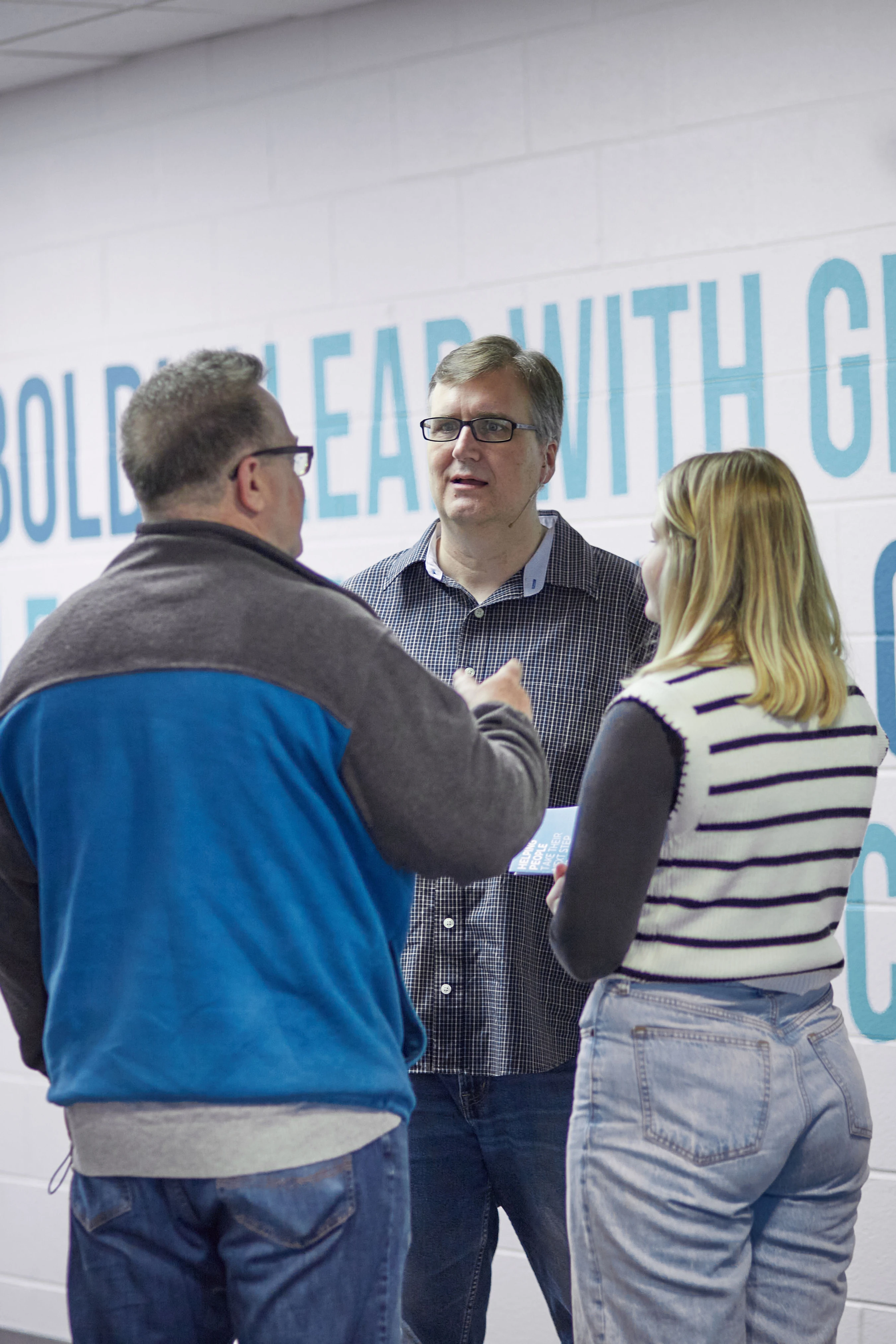 A man with glasses stands between another man and a woman as they talk. They are in a hallway with large, light blue motivational text on the white wall.