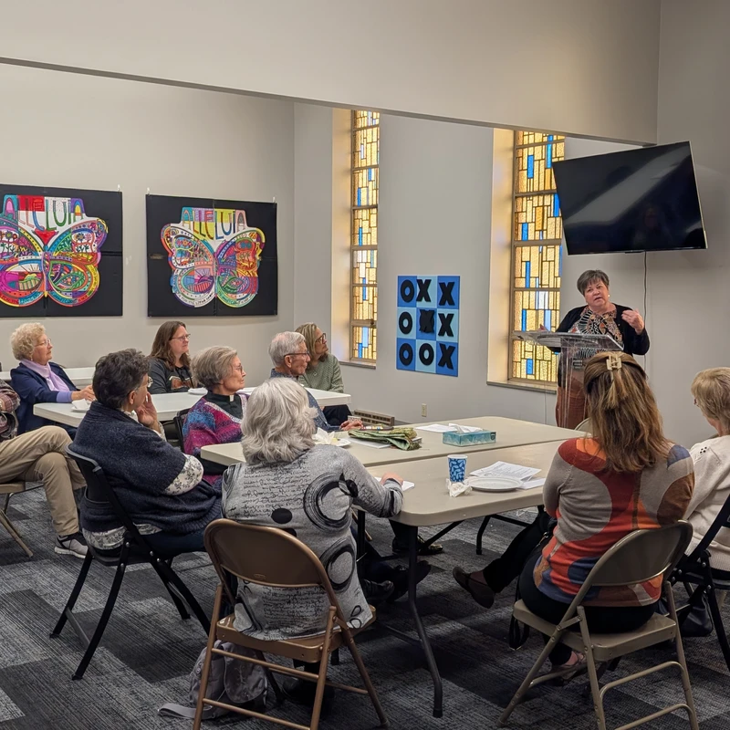 A woman at a podium speaks to a group of mostly older adults seated at tables. The room has stained glass windows and colorful butterfly artwork on the wall.