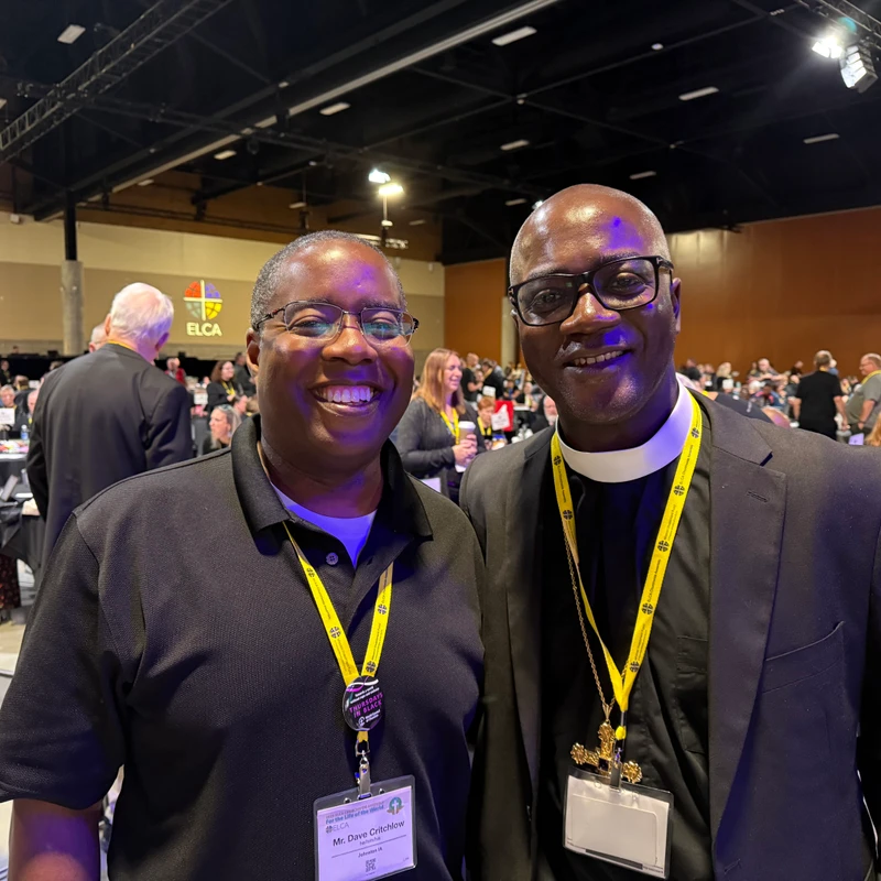 Two Black men, one a pastor in a clerical collar, smile for the camera while standing in a large hall during an ELCA church assembly.