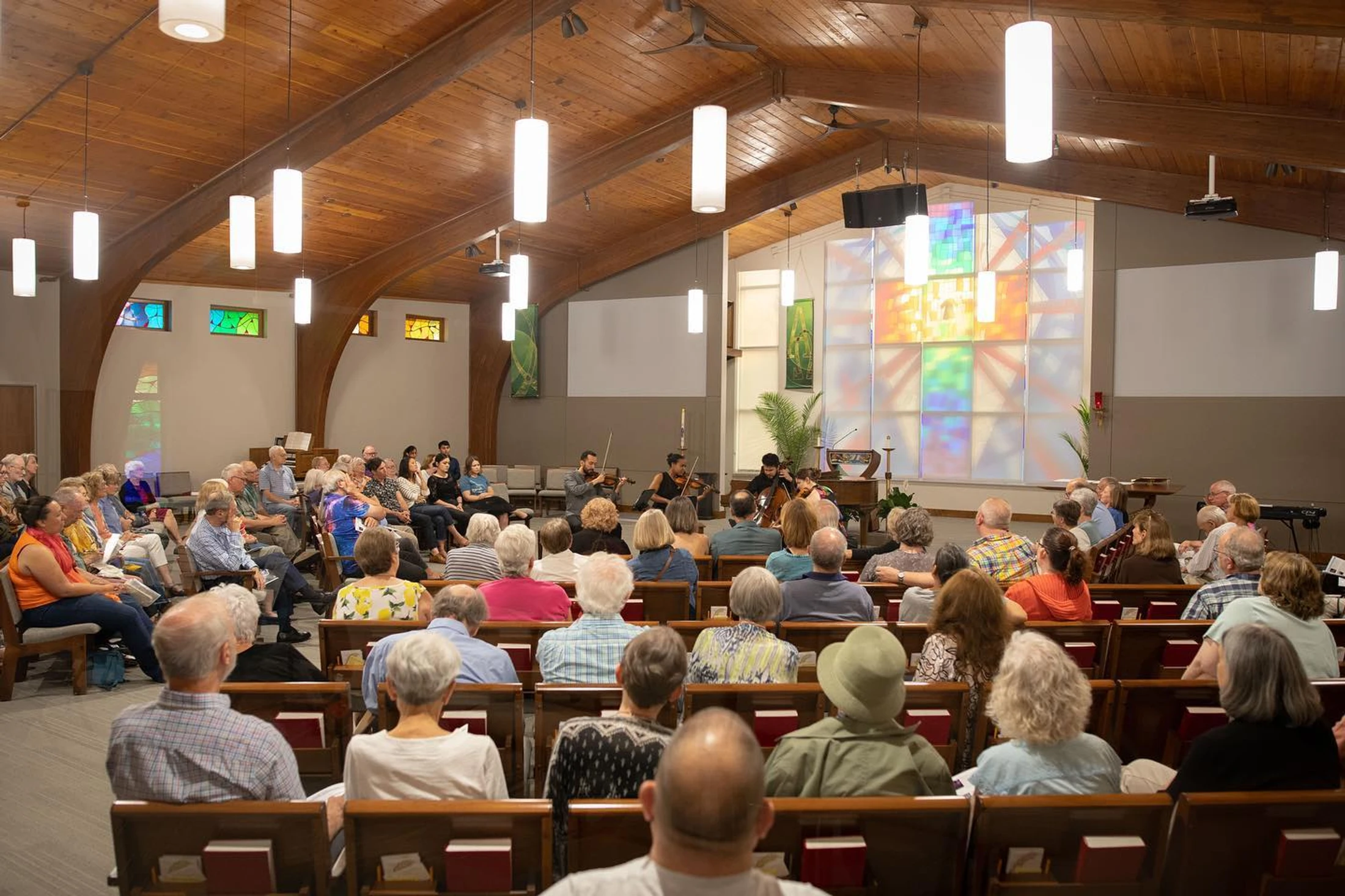 An audience sits in pews watching a string quartet perform in a modern church with a high, wood-paneled ceiling and a large, colorful window.