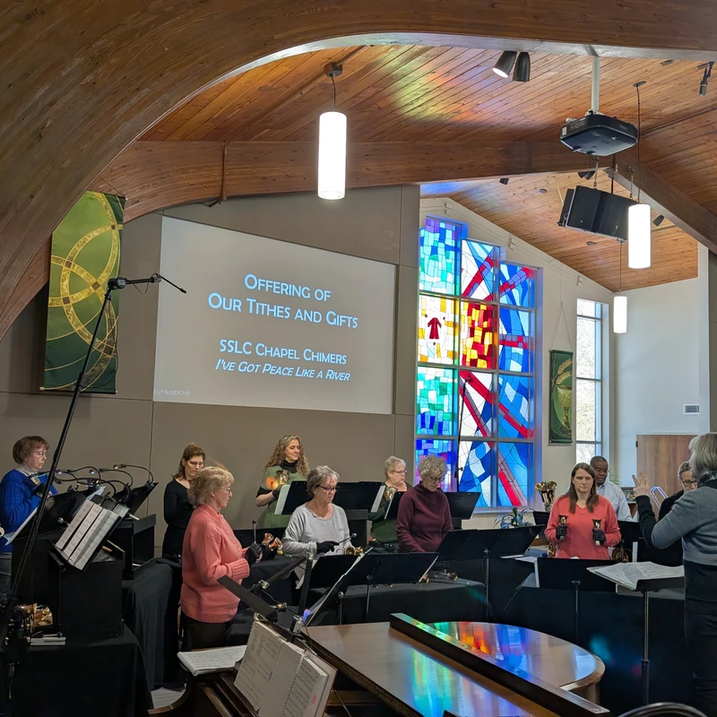 A chime choir performs in a modern church sanctuary with a curved wooden ceiling and stained glass windows, directed by a conductor.