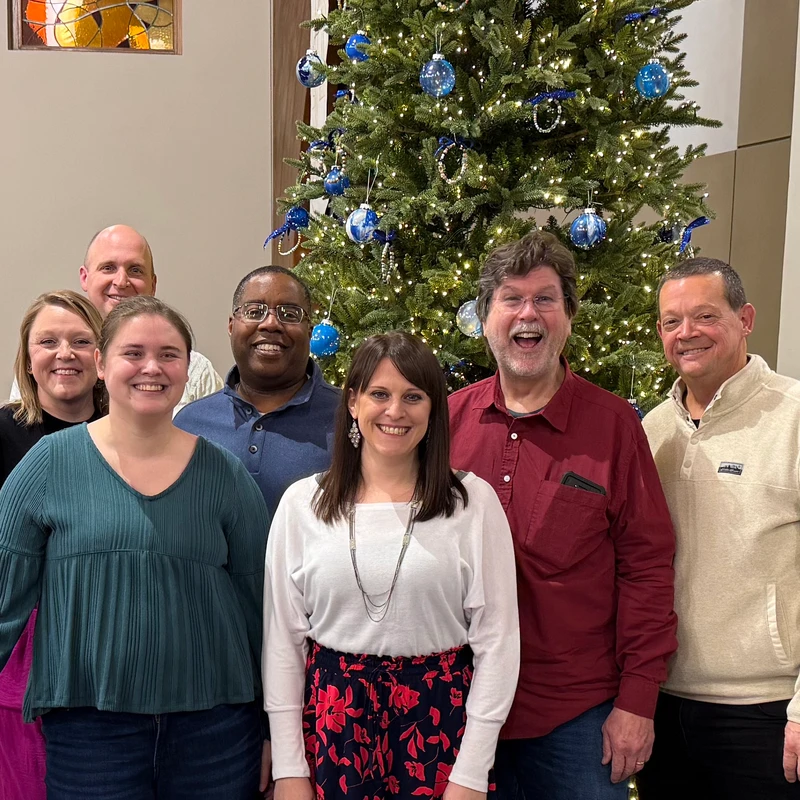 A group of seven smiling adults stand together for a photo in front of a Christmas tree decorated with blue ornaments and white lights.
