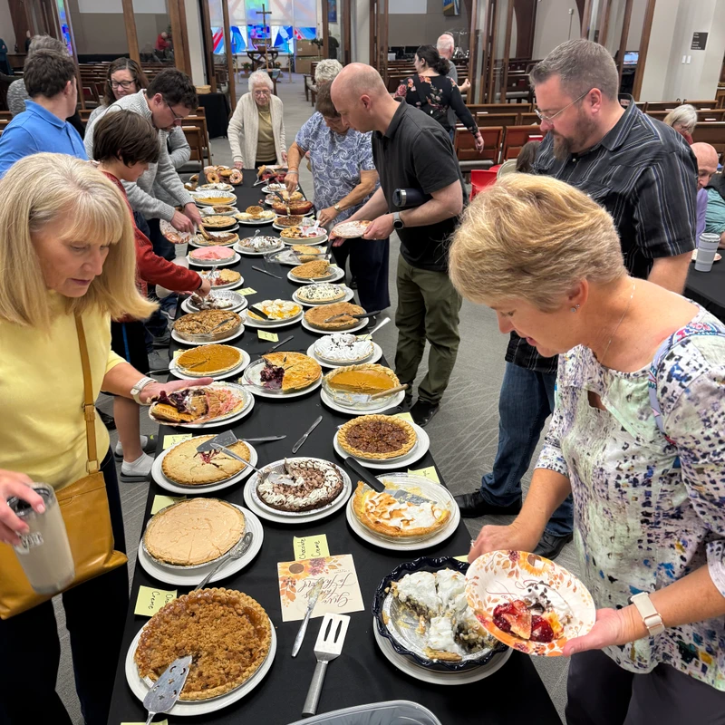 A group of people standing around a long table covered in dozens of different pies, serving themselves slices onto paper plates at a social event.