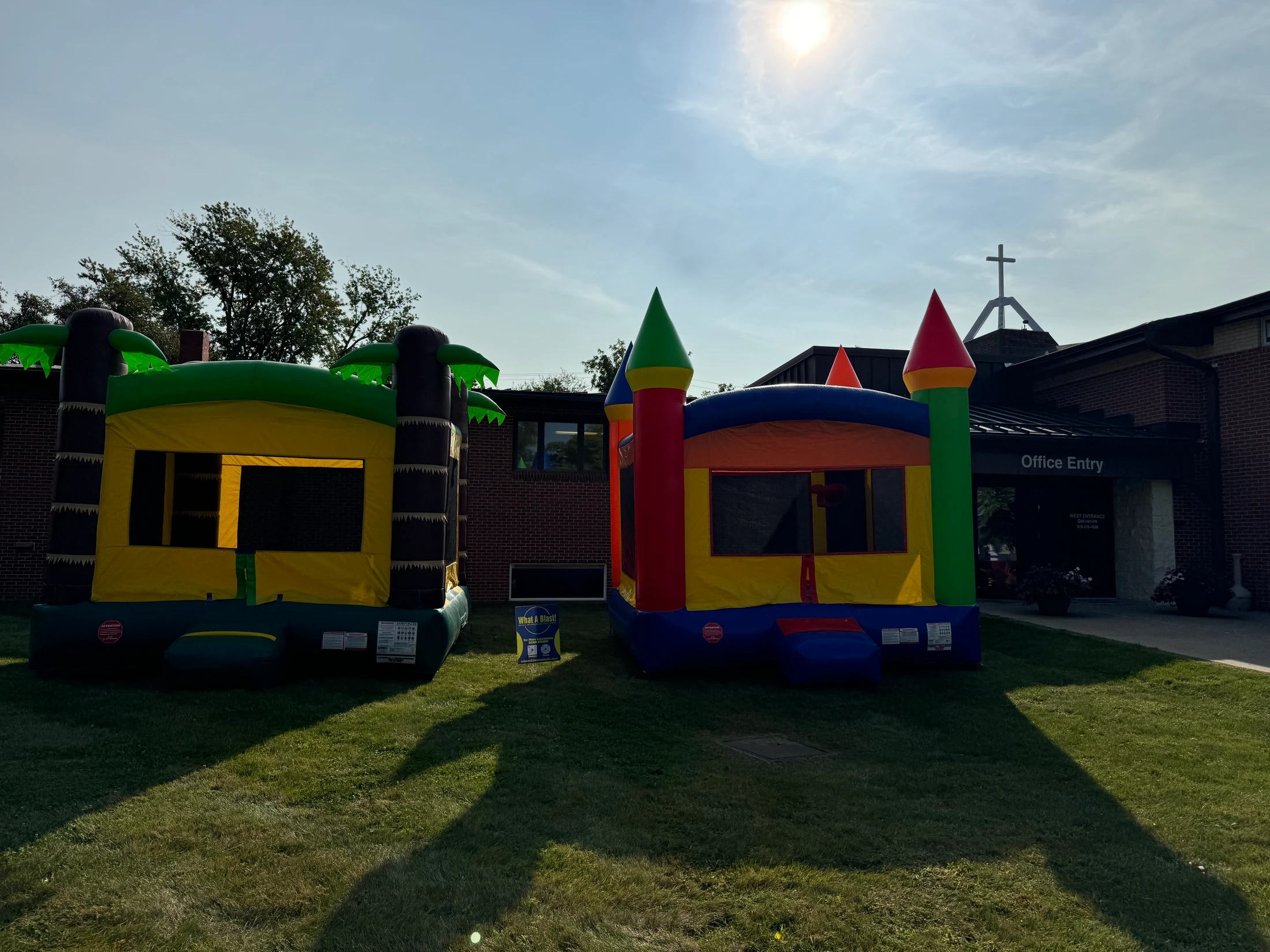 Two colorful bounce houses, one castle-themed and one palm tree-themed, sit on a grassy lawn in front of a brick church building on a sunny day.
