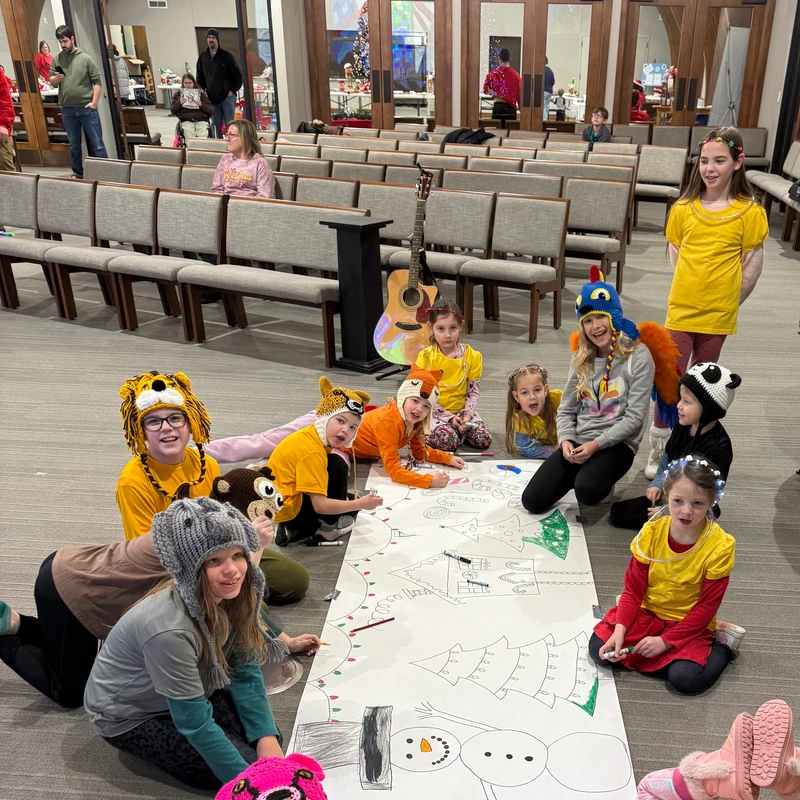 A group of children wearing funny knitted animal hats sit on the floor of a large room, coloring a long paper banner with a Christmas scene.