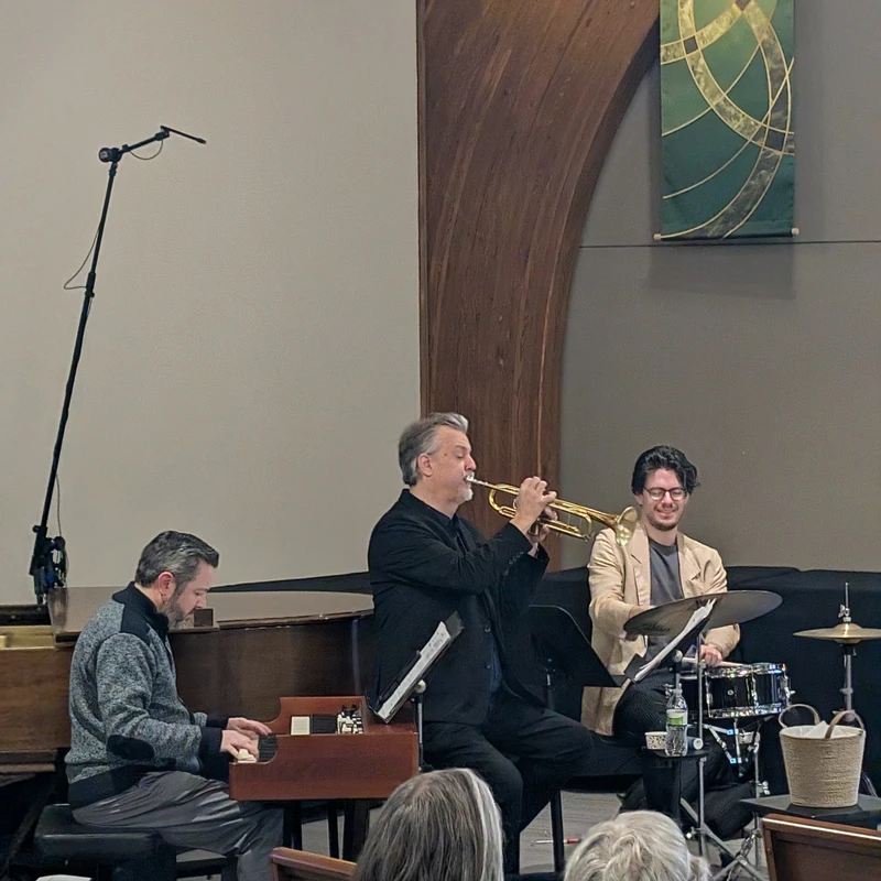 A jazz trio with a pianist, a trumpeter, and a drummer performs on a stage in a church for an audience seated in wooden pews.