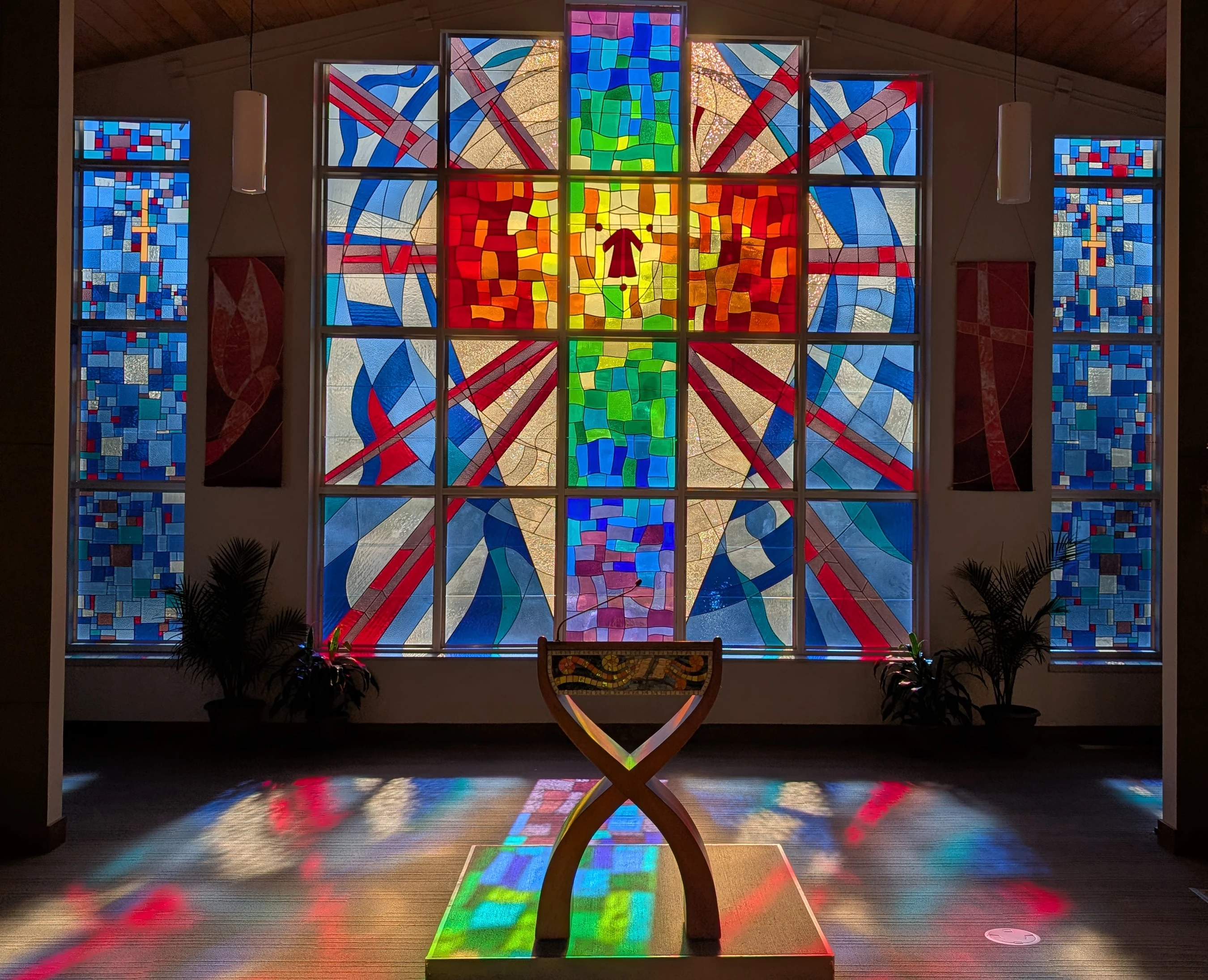 A large, modern stained glass window with a rainbow-colored cross design illuminates a dim chapel, casting vibrant colors on the floor and a wooden stand.