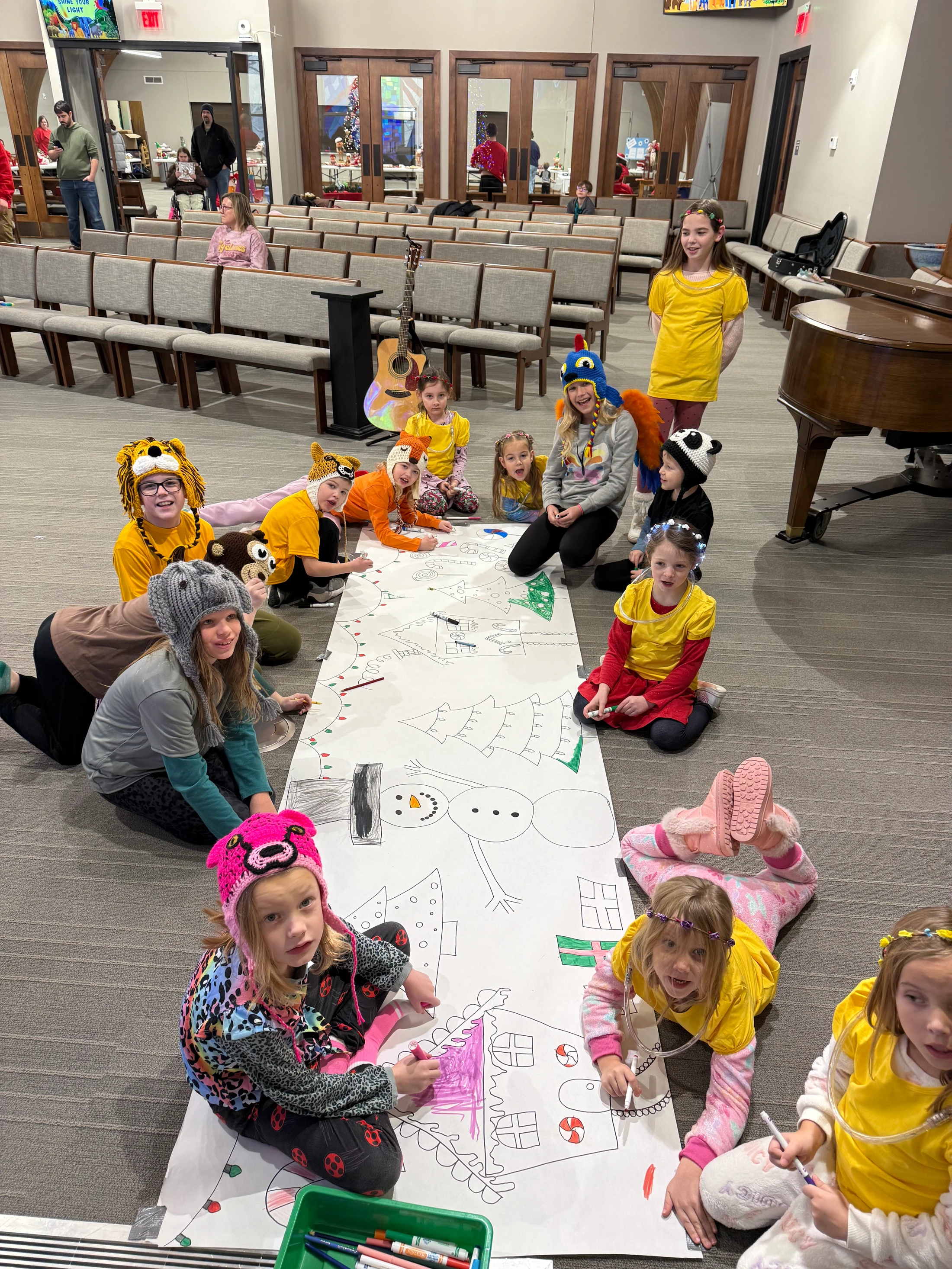 A group of children wearing silly animal hats sit on the floor coloring a giant holiday-themed banner with markers in a large hall.