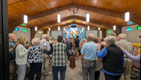 A congregation stands in a circle inside a church with a high wooden ceiling, placing hands on each other's shoulders in a moment of community prayer.