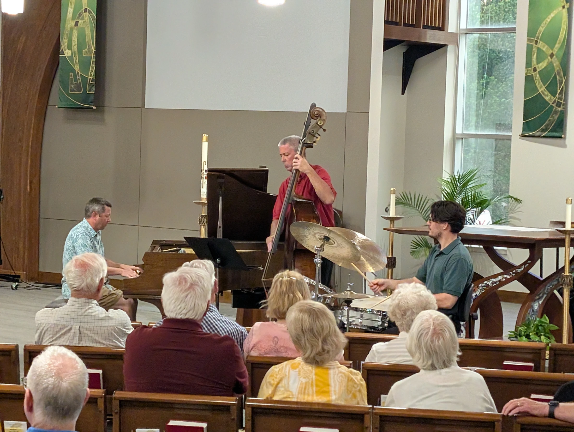 A jazz trio with a pianist, double bassist, and drummer performs for an audience seated in wooden pews inside a modern-looking church.
