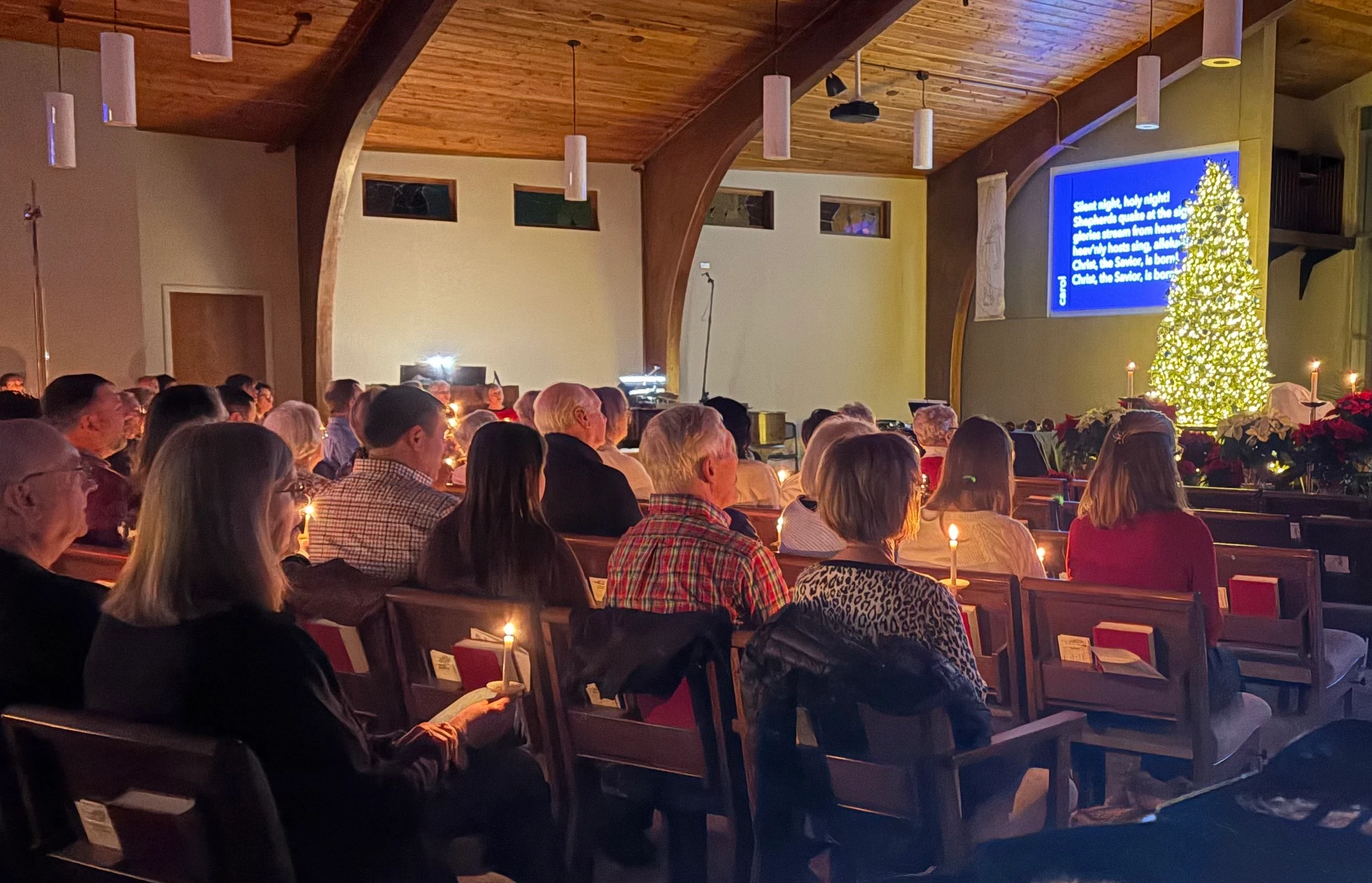A congregation seen from behind sits in church pews, holding lit candles during a Christmas service. A lit Christmas tree is at the front.