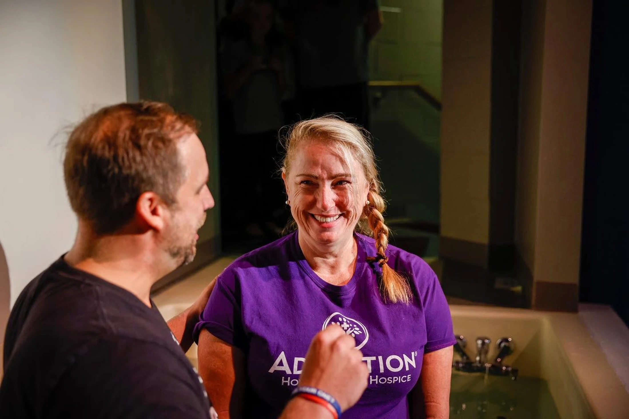A smiling blonde woman in a purple t-shirt stands in a baptismal font, looking at the camera, as a man in a black shirt stands beside her.