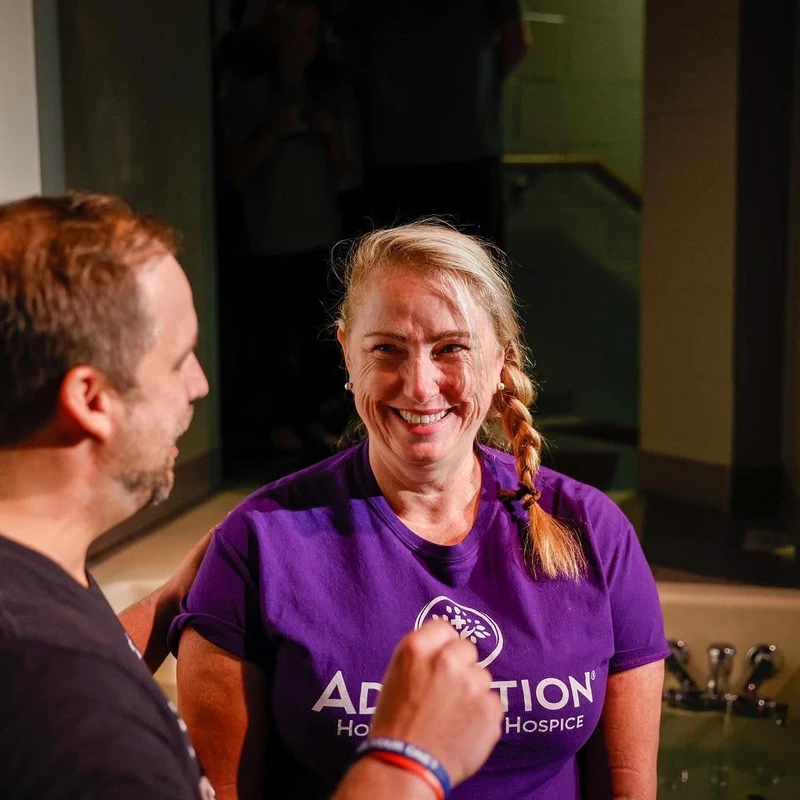 A smiling blonde woman in a purple t-shirt stands in a baptismal font, looking at the camera, as a man in a black shirt stands beside her.