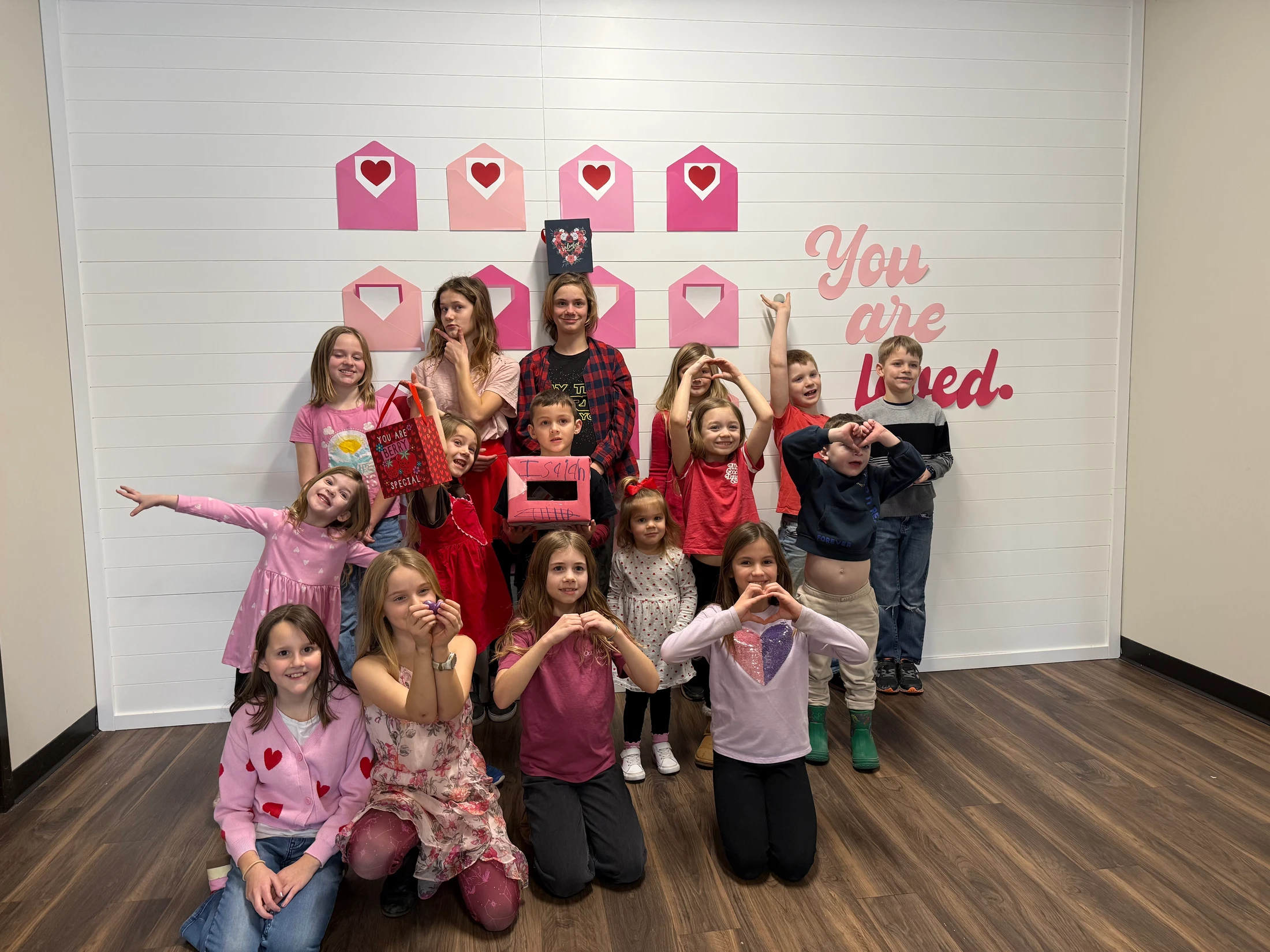 A group of children and a teenager pose for a photo in front of a wall with pink hearts and the words 'You are loved' for a Valentine's Day celebration.