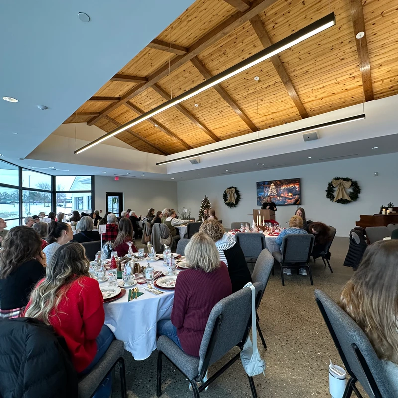 A group of women seated at round tables listen to a speaker at a podium during a Christmas luncheon in a spacious room with a vaulted wood ceiling.