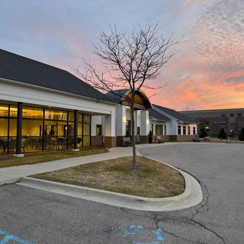 A modern white building with large, glowing windows stands next to a parking lot. A bare tree is silhouetted against a beautiful, colorful sunset sky.