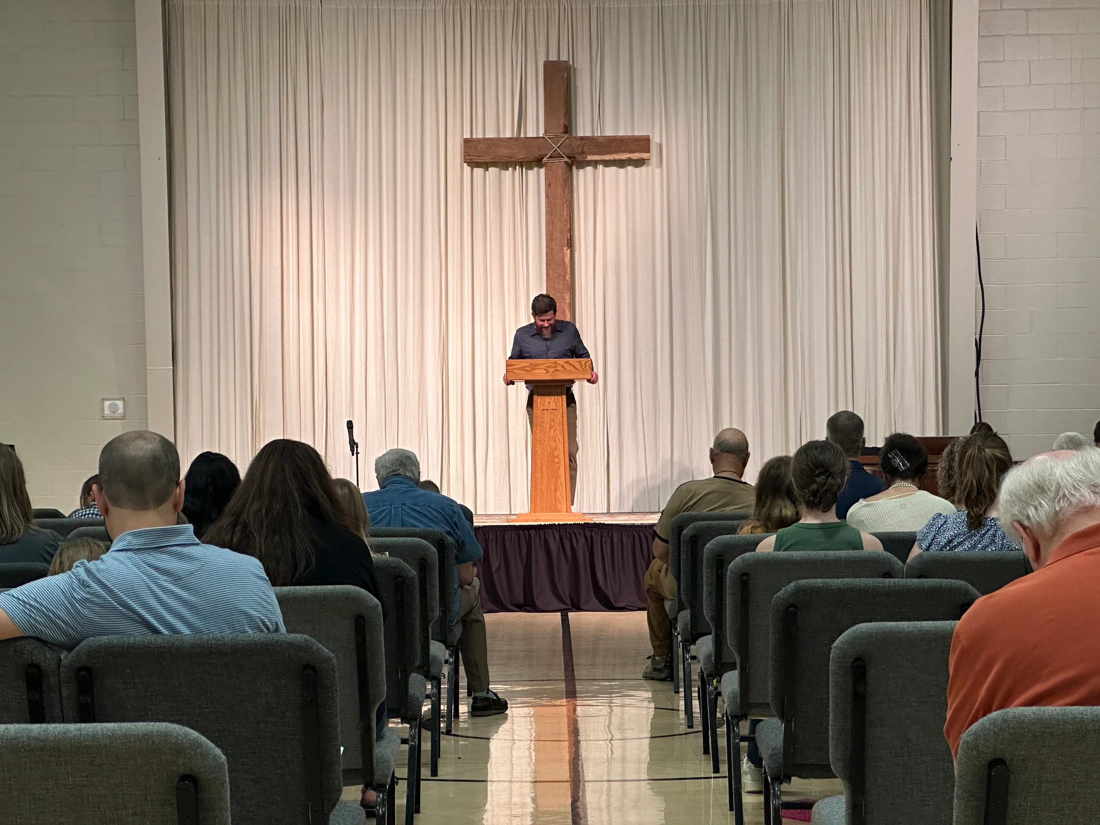 View from the back of a seated congregation as a pastor preaches from a wooden pulpit. A large wooden cross is on the stage behind him.