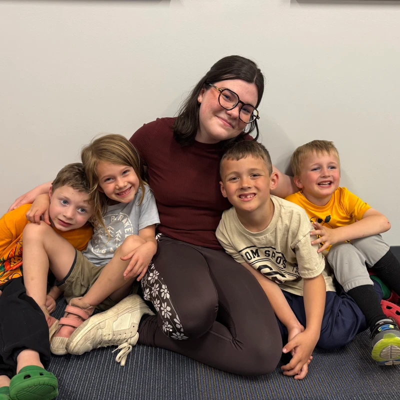 A woman with glasses and four young children sit together on a blue carpeted floor, smiling and posing happily for a group picture against a plain wall.