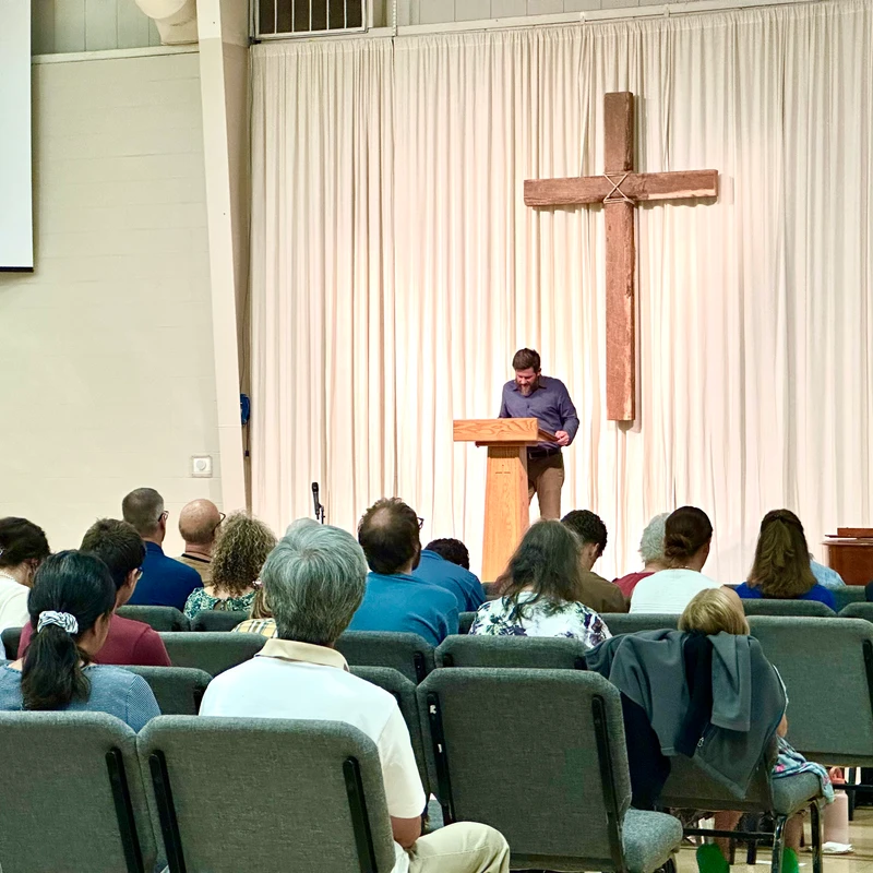 A view from the back of a congregation as a pastor preaches from a wooden lectern on a stage. A large wooden cross hangs on the curtain behind him.