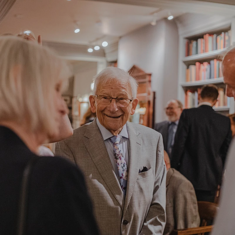 An elderly man with white hair and glasses, wearing a grey suit and floral tie, smiles warmly while mingling with guests at an indoor social event.