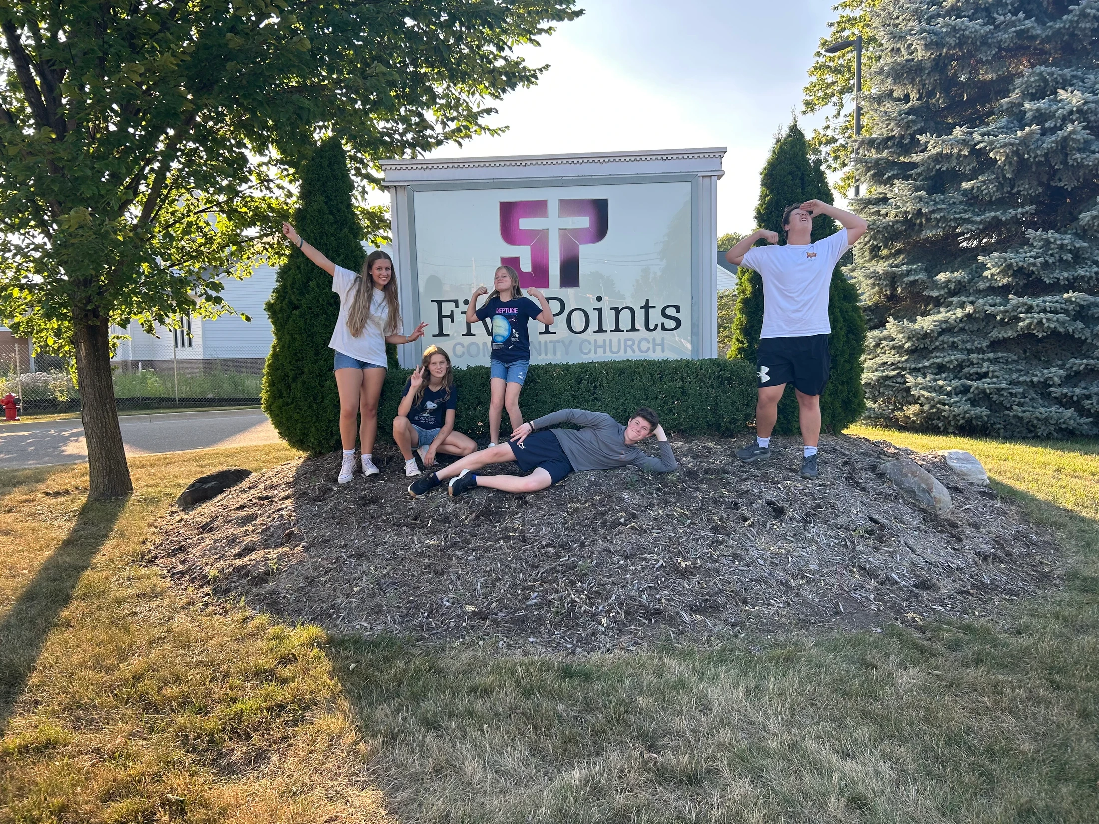 A group of five young people making funny and playful poses for a photo in front of the Five Points Community Church sign on a sunny day.