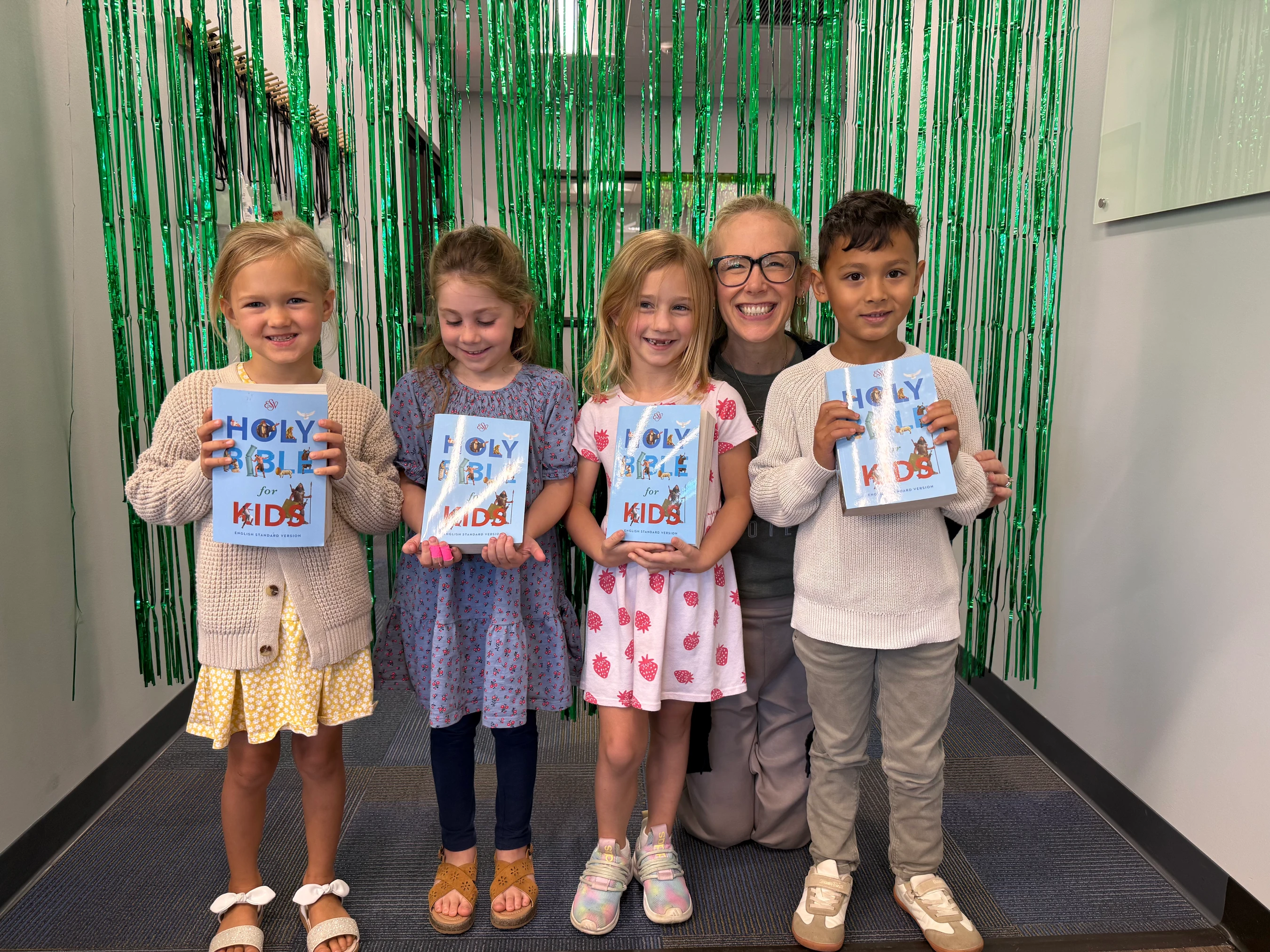A woman and four young children stand in front of a green curtain, all smiling and holding copies of the 'Holy Bible for Kids'.