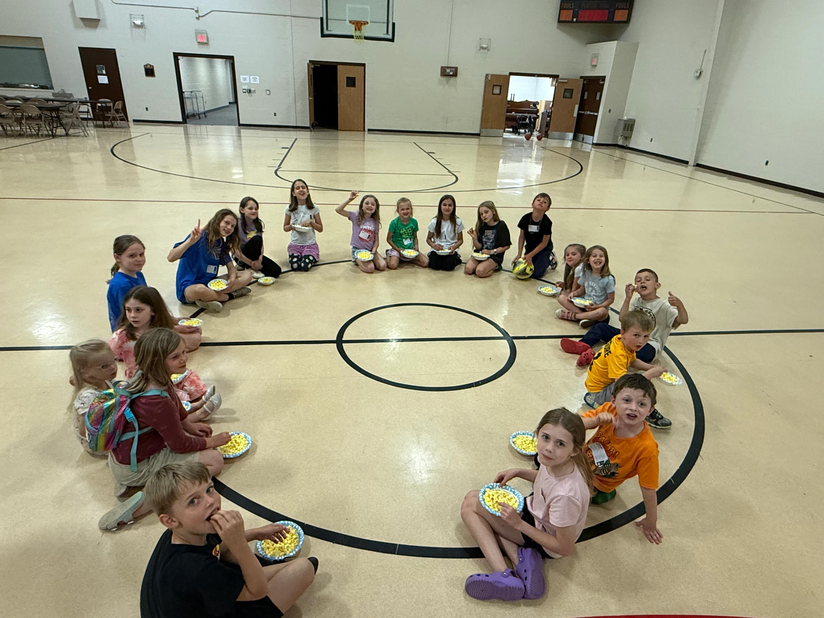 A large group of elementary school-aged children sit in a circle on a gymnasium floor, smiling and eating popcorn from paper plates.