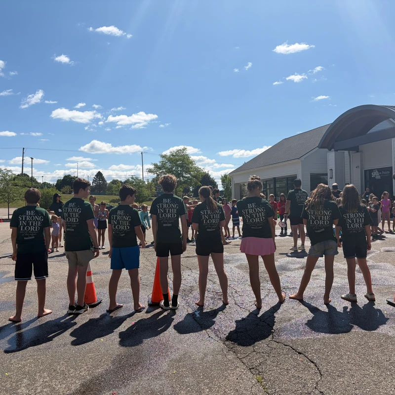 A group of teens stands in a line with their backs to the camera, wearing matching green shirts that say 'Be Strong in the Lord' in a sunny parking lot.