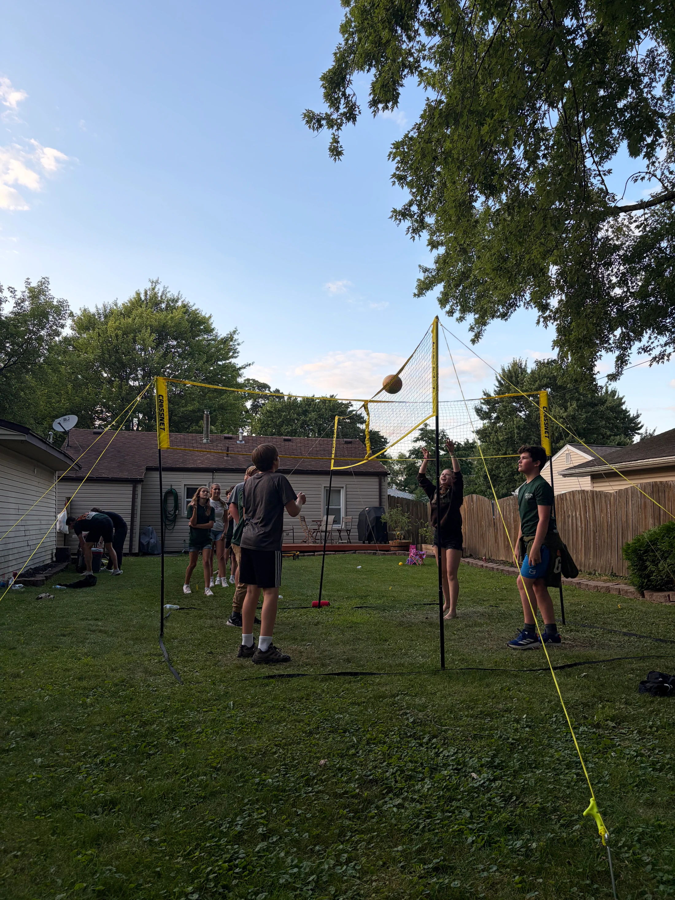 A group of young people play a game of four-way volleyball in a grassy backyard as the sun sets, with one girl hitting the ball over the net.