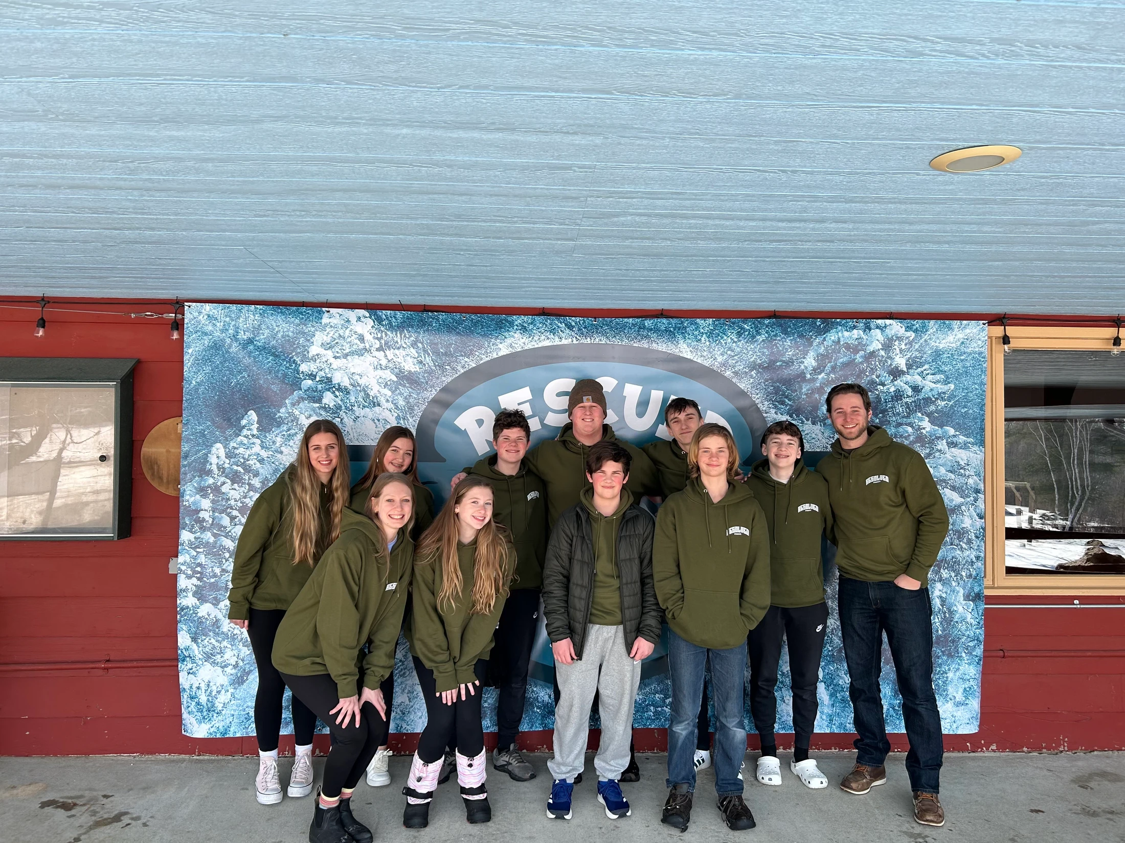 A group of eleven young people and one adult pose for a photo in matching olive green hoodies in front of a blue and white winter-themed banner.