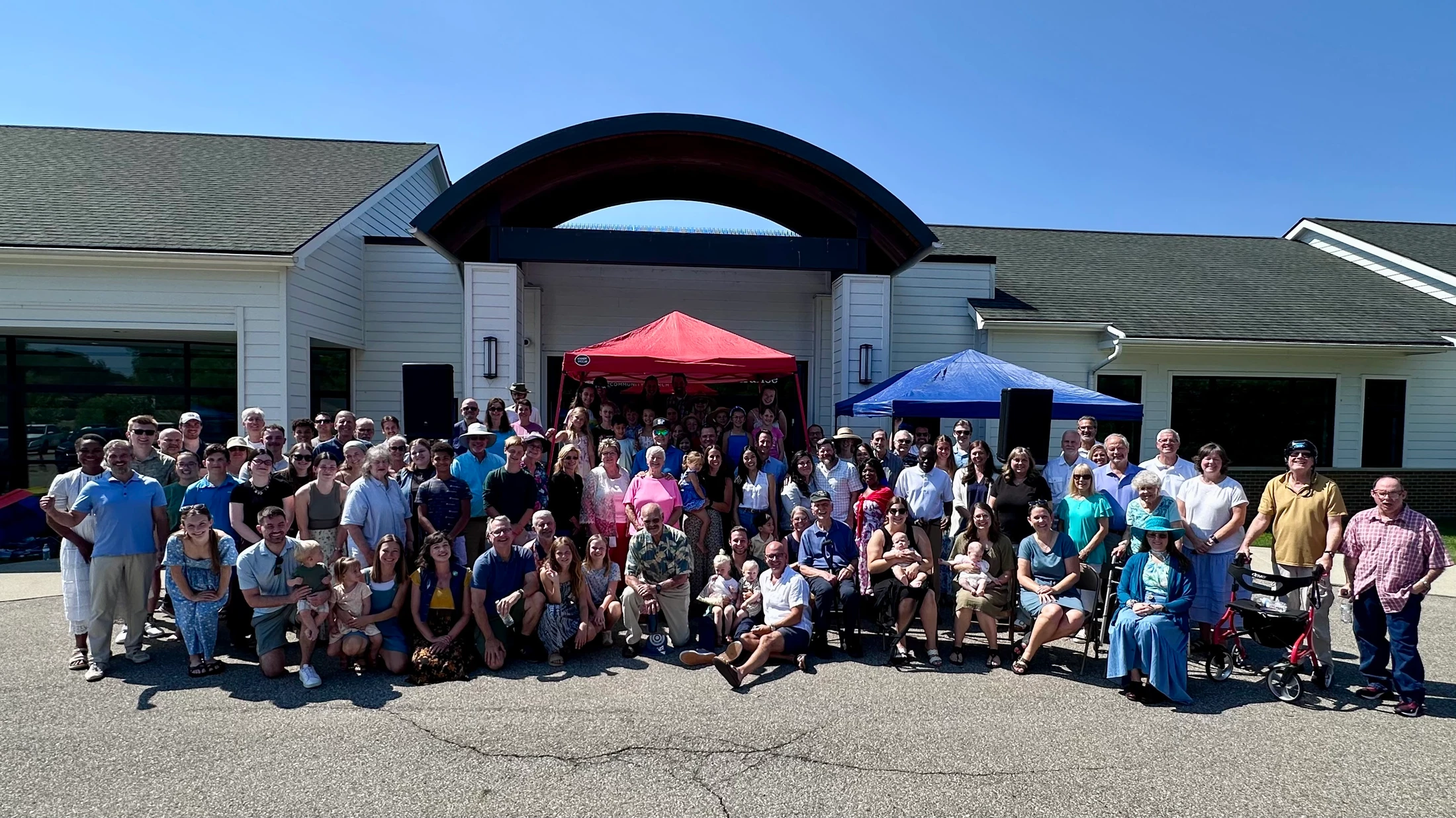 A large, diverse group of people of all ages posing together for a photograph outdoors in front of a modern building under a clear blue sky.