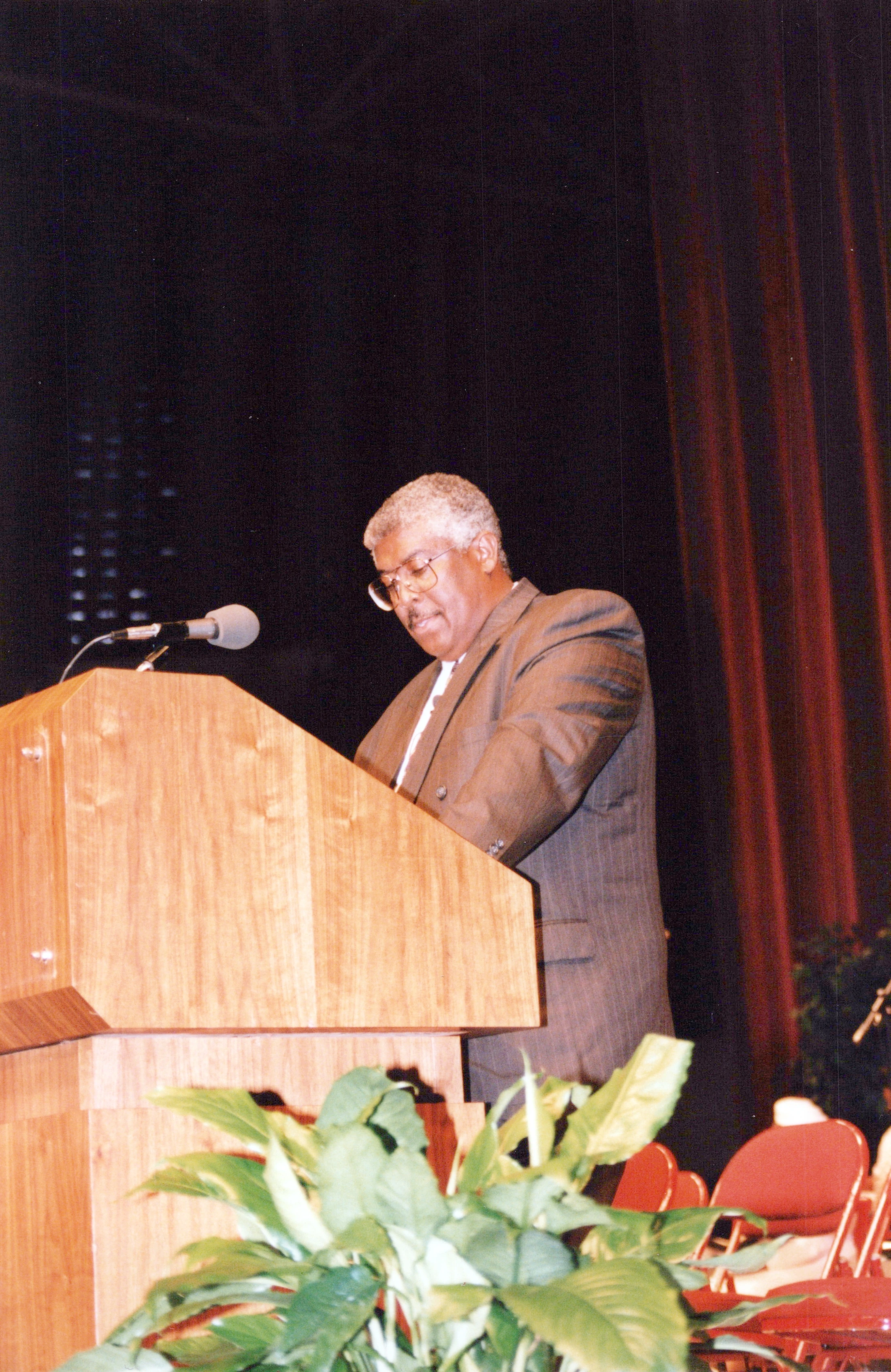 An older Black man with gray hair and glasses wears a pinstripe suit while standing at a wooden podium on a stage, looking down as if reading a speech.