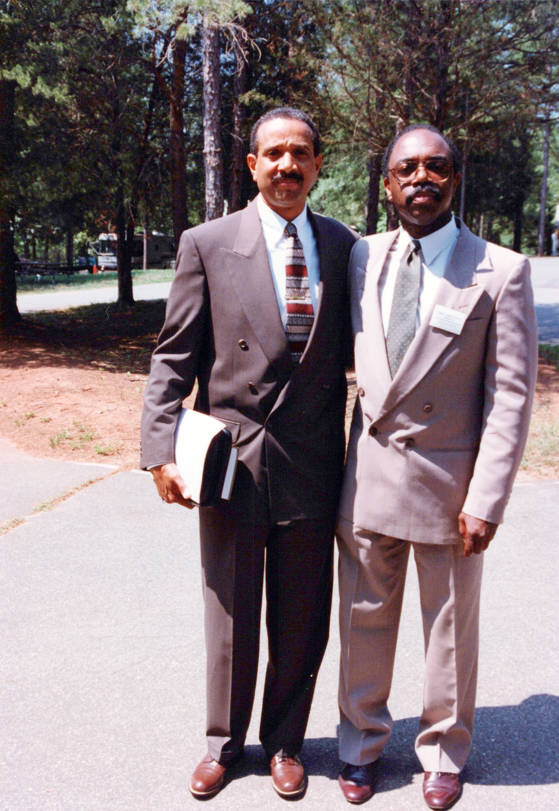 A full-length outdoor portrait of two Black men in double-breasted suits. They stand side-by-side on a paved path with trees in the background.