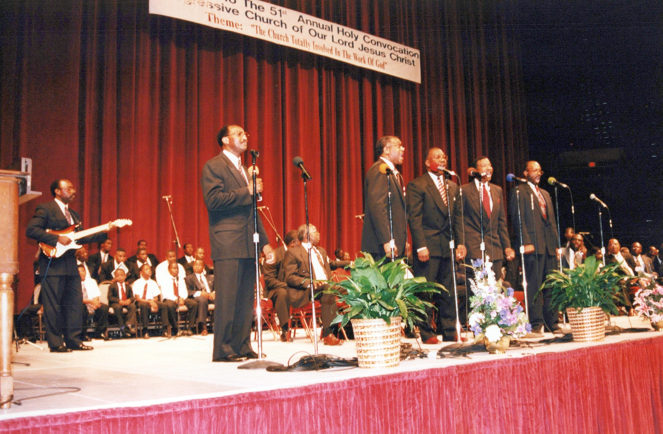 A group of African American men in suits sing on a stage with a red curtain. One man plays guitar while others sing into microphones at a church convocation.