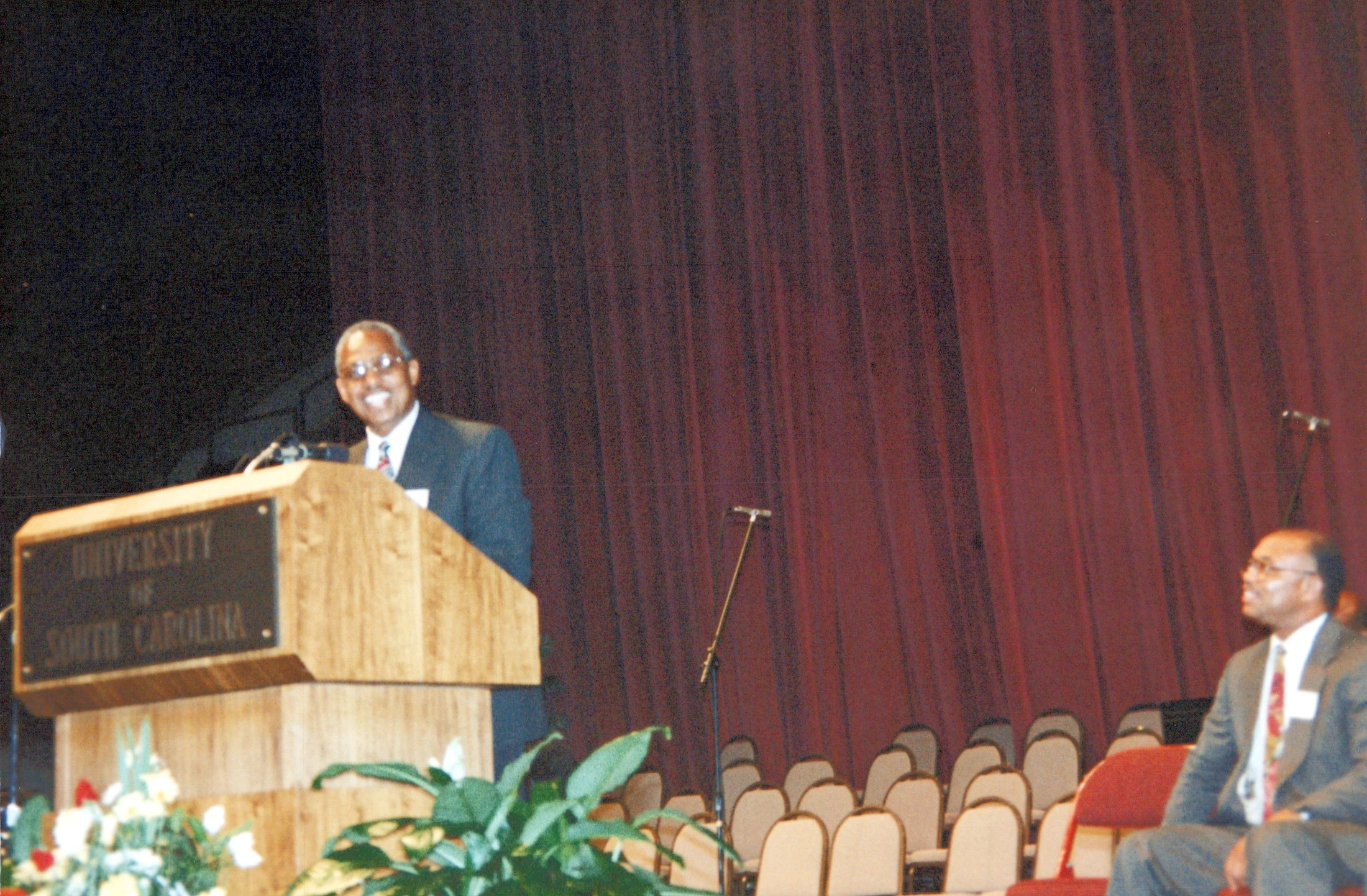 A smiling man in a suit and glasses speaks at a wooden podium with a "University of South Carolina" sign. Another man sits to the side watching.
