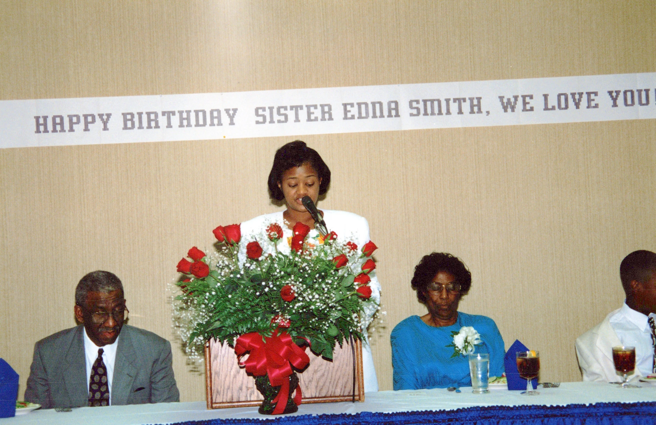 A woman speaks into a microphone at a party. Behind her, a banner reads 'Happy Birthday Sister Edna Smith.' Guests sit at a table with a large floral display.