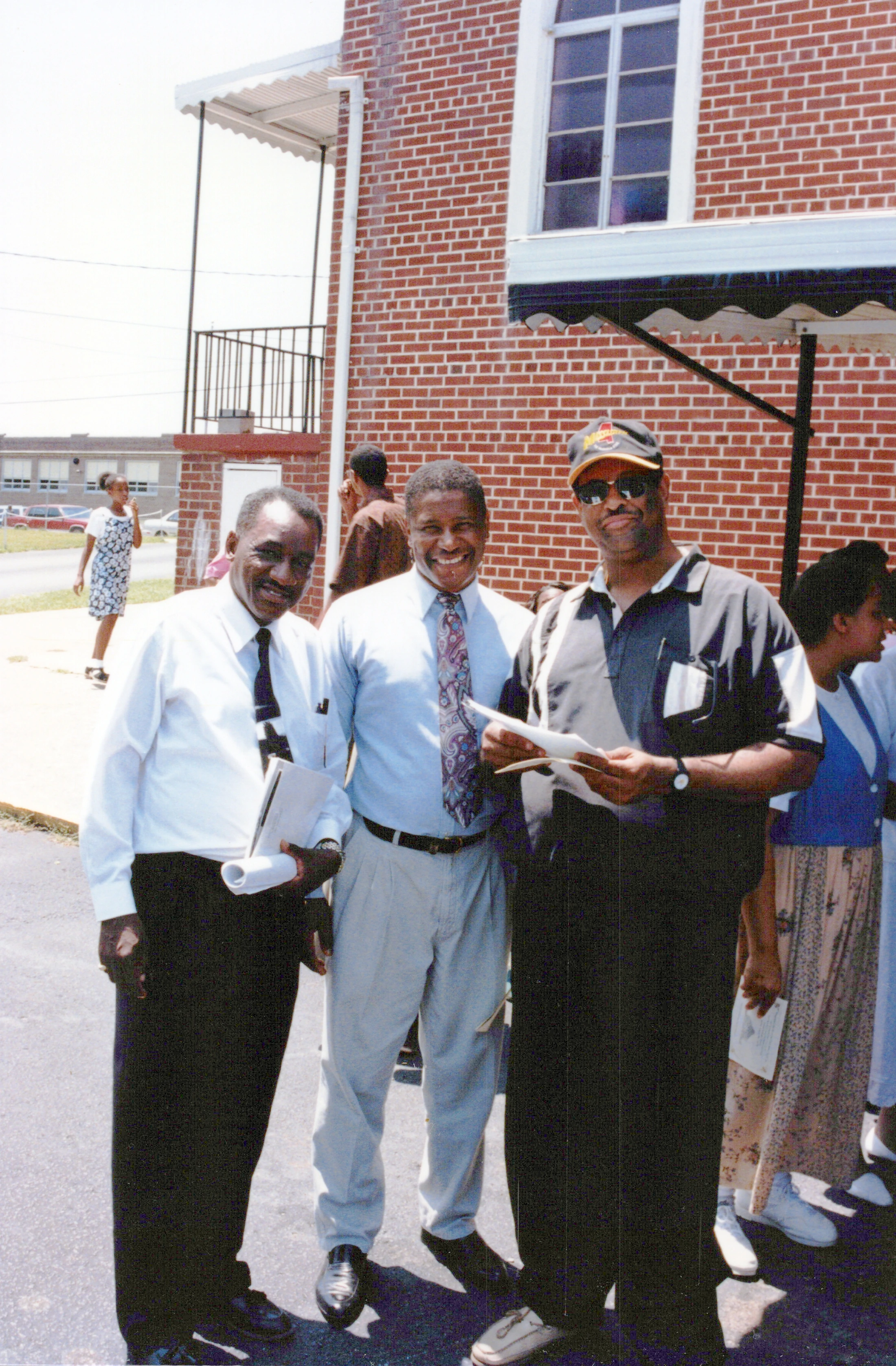 Three smiling African American men stand together for a photo outside a red brick building on a sunny day. Other people are in the background.