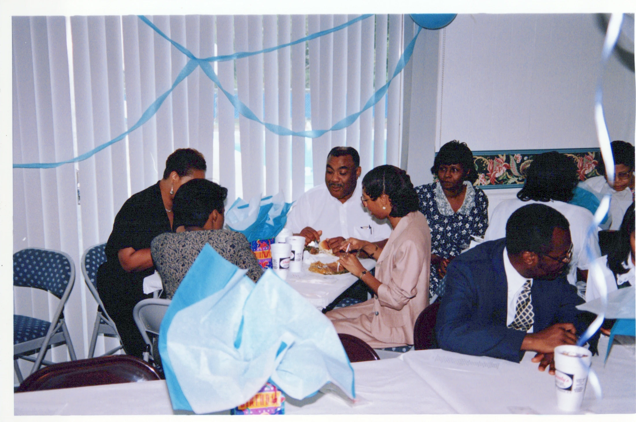 A vintage photo shows a group of Black adults sitting at tables, eating and socializing at an indoor party decorated with blue streamers.