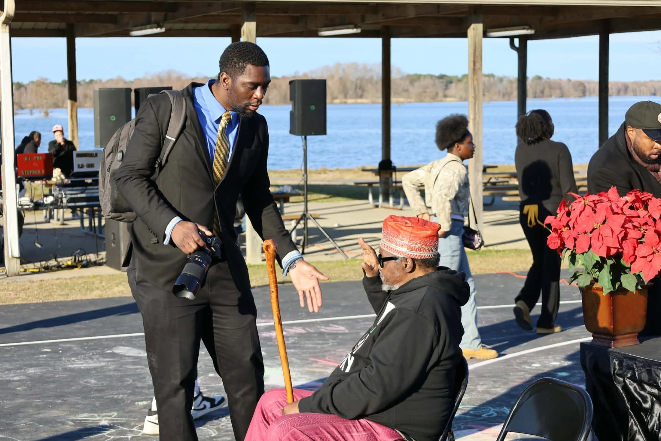 A young Black man in a suit with a camera leans down to speak with a seated older Black man in a red hat who gestures with one hand while holding a cane.