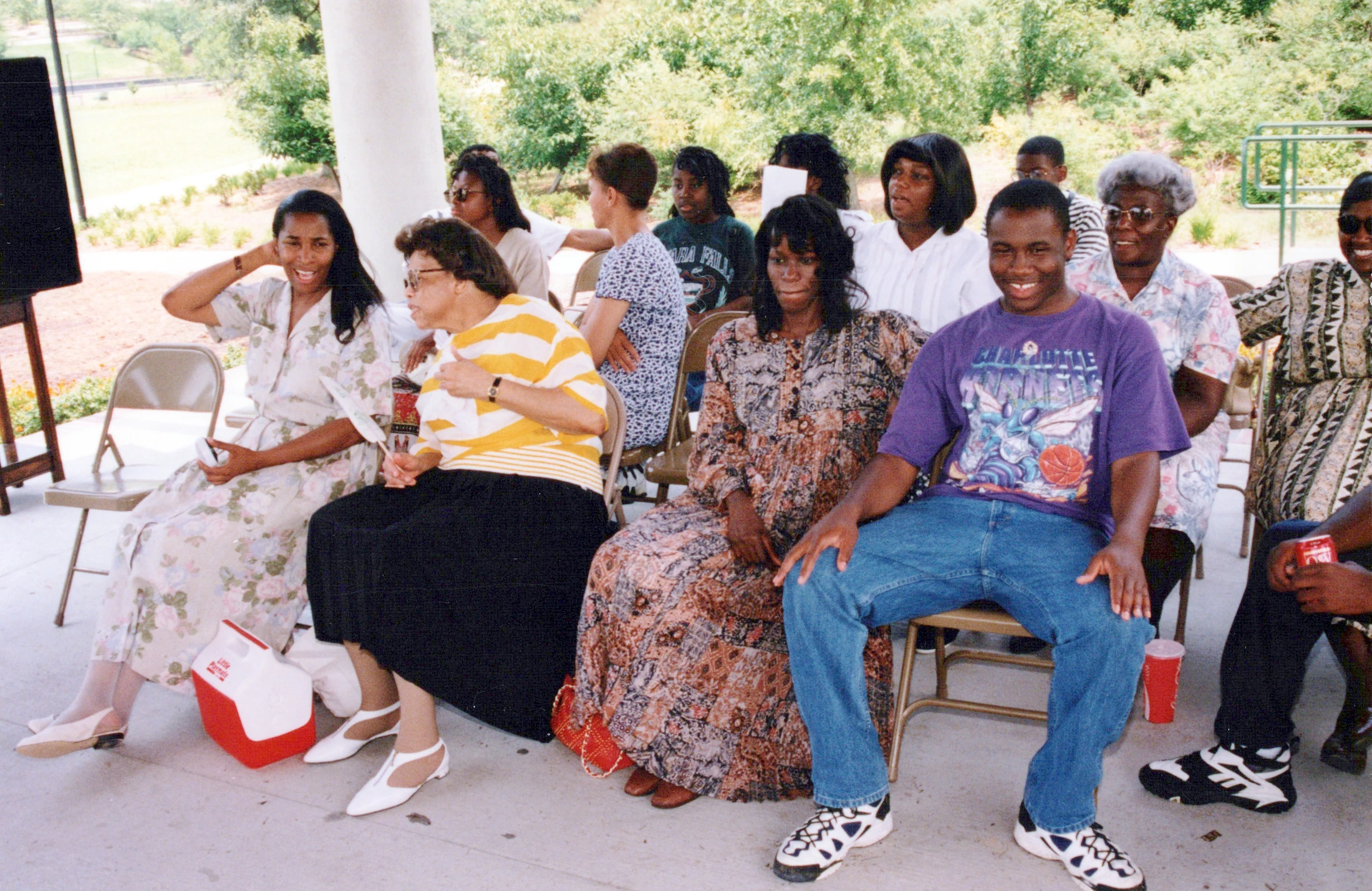 A group of men and women, mostly African American, sit in folding chairs under a pavilion, smiling and looking towards the camera at a daytime event.